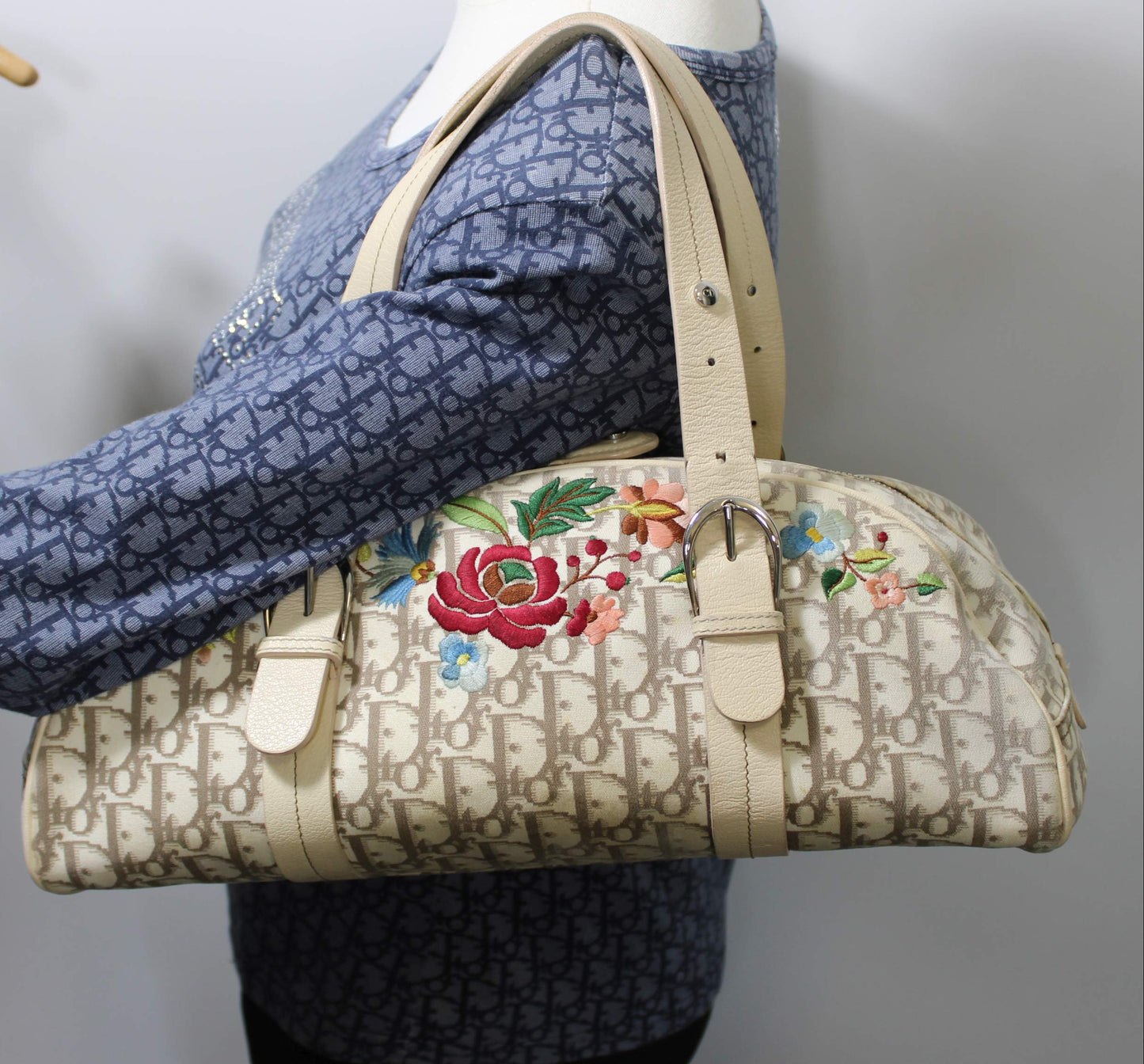 A beige monogram handbag with colorful floral embroidery on the front, displayed on a mannequin wearing a blue patterned top against a plain light gray background.